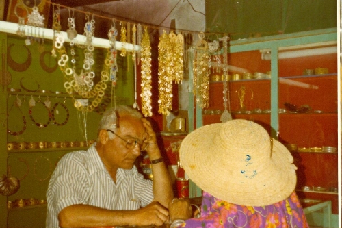 Sheikh Ouzifa Houri (bijoutier), commerçant dans sa boutique avec une cliente de dos avec un chapeau de paille.